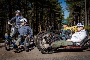 Four people using adaptive bikes and all terrain wheelchairs on forest track.