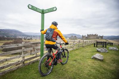 A cyclist leaning against a sign with Ruthven Barracks in the distance.