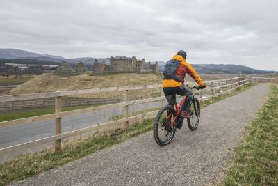 Person riding a bike on the Speyside Way path with Ruthven Barracks in the background.