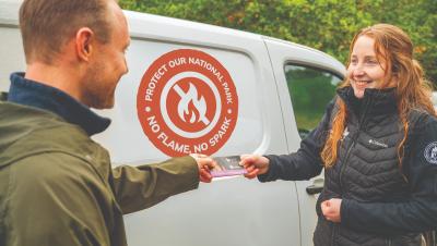 A Park Authority ranger hands out a fire leaflet to a member of the public, in front of a white Park Authority ranger van with an orange 'No flame, no spark' graphic on the side.