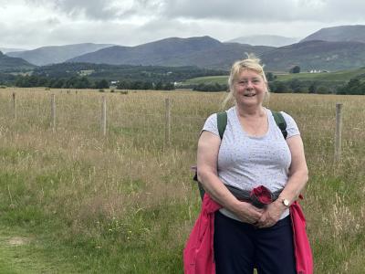 Caron Dawson standing smiling on a path at Kingussie Dell, with a field and hills in the background.