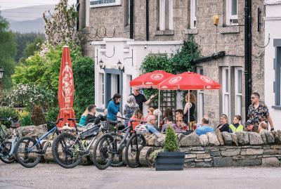 People sitting and standing in a hotel beer garden with red parasols above their heads and bikes in the foreground.