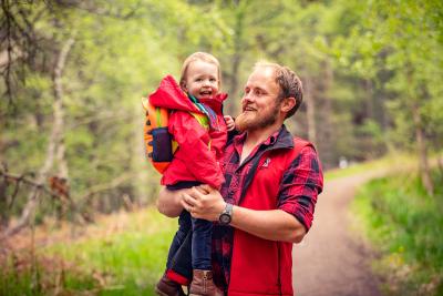 An adult holding a child both smiling on a path with trees in the background