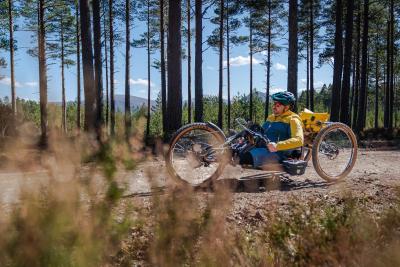 A person using an adaptive bike on forest tracks with trees and hills in the background.