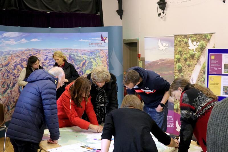 Group of six people stood around a table with a map on it, in deep discussion. Behind them are two Park Authority staff stood in front of a large map of the National Park, as well as two pop-up banners to their right.