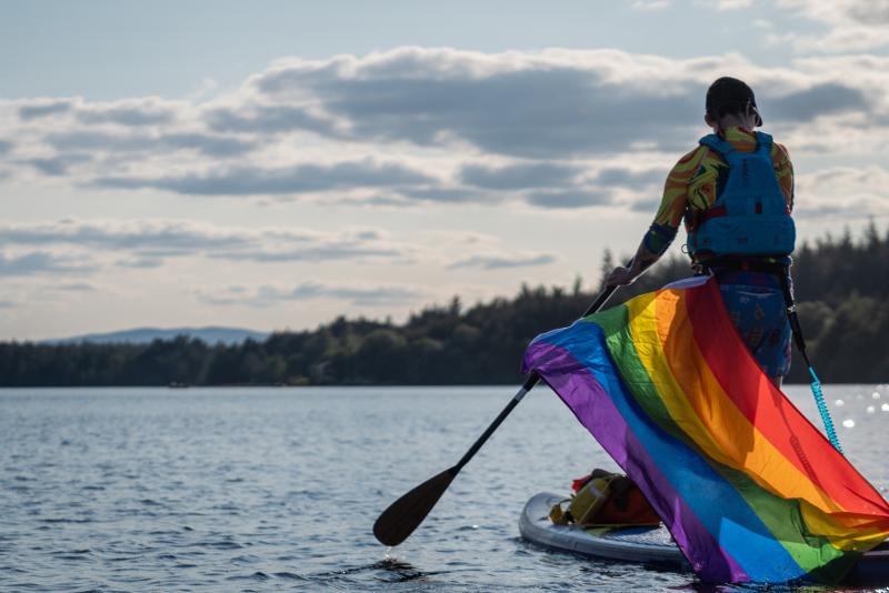 A person paddleboarding on Loch Morlich with an LGBTQ+ flag