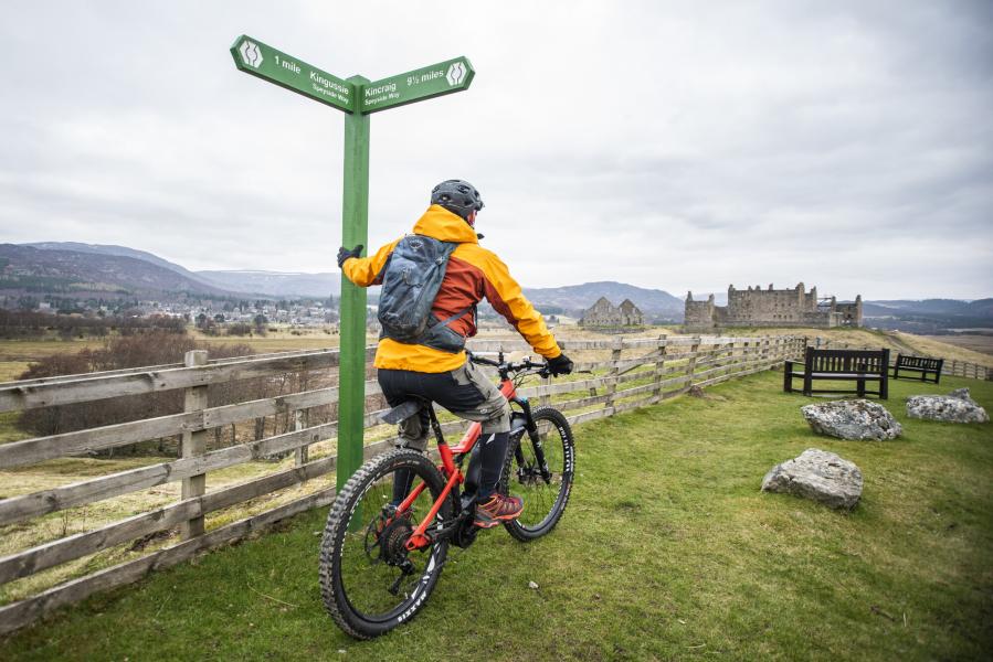 A cyclist leaning against a sign with Ruthven Barracks in the distance.