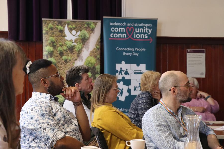 Seven people sit listening to a talk with banners in the background