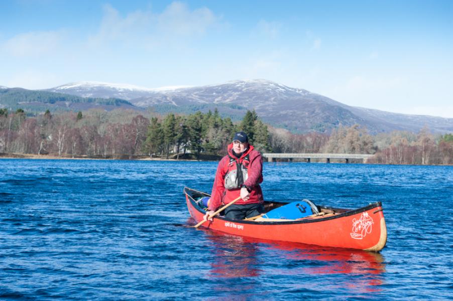 A person in an open canoe on Loch Insh with bridge and mountain in the background.