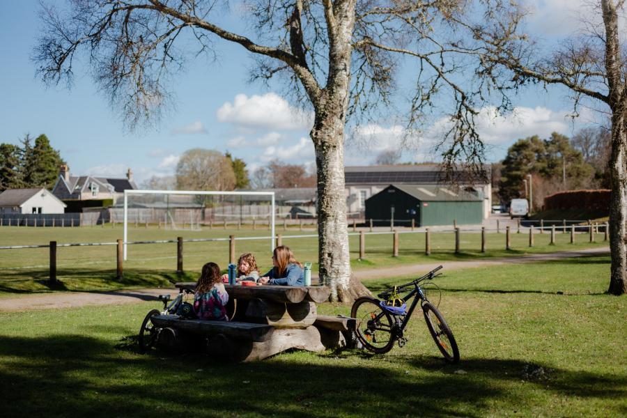 A family sits at an outdoor picnic bench. There is a playing field, football goals, and a tree in the background. The children's bikes are propped up against the wooden picnic bench.