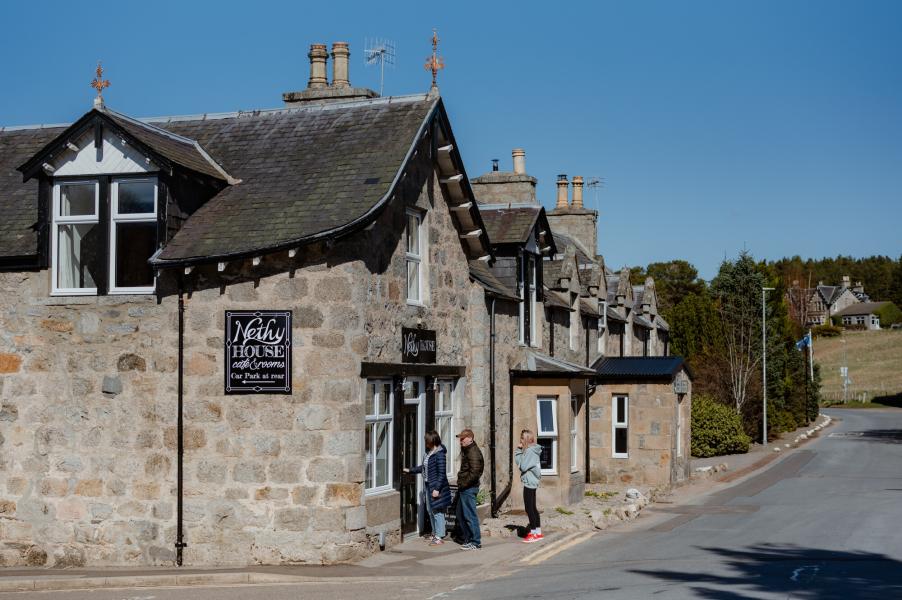Two adults and a teenager go into a shop in Nethy Bridge. The shop is on the corner of the street, and the building is grey coloured stone with a black slate roof and several chimneys.