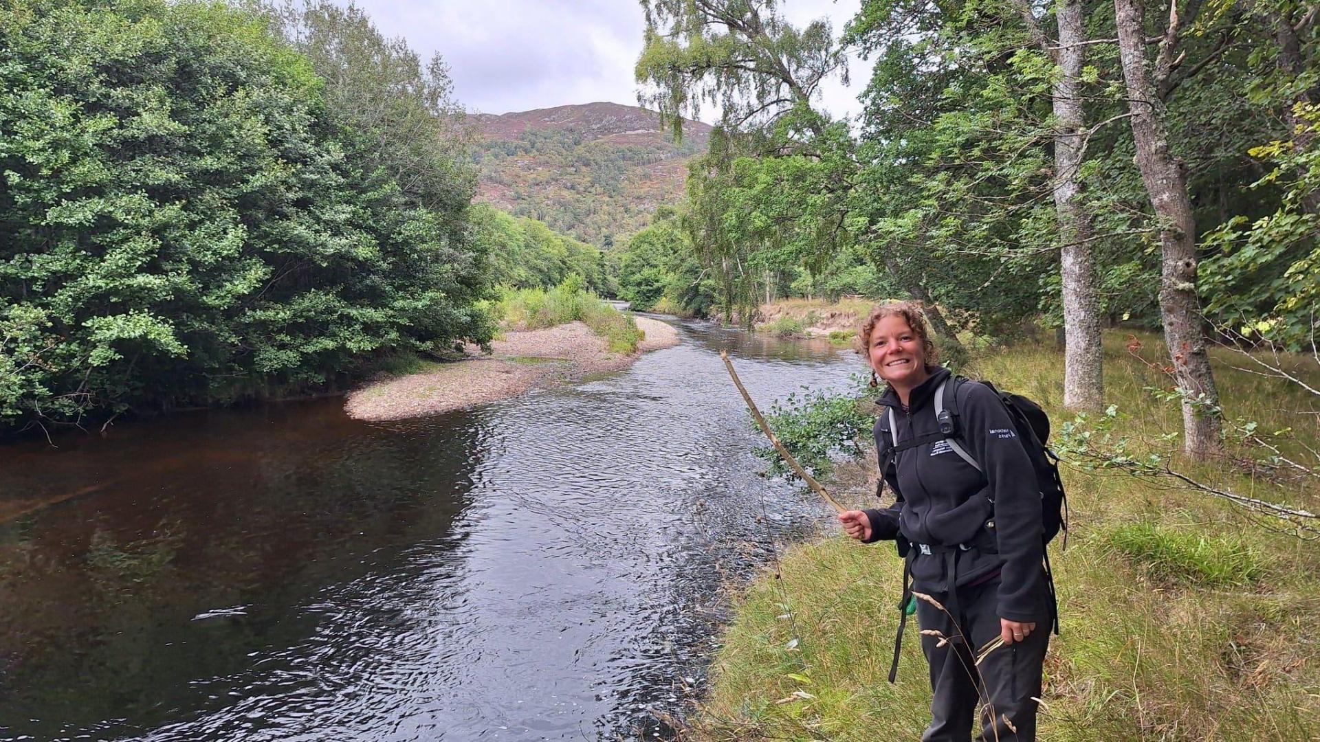 Georgia smiles wearing a rucksack in front of a river and hill
