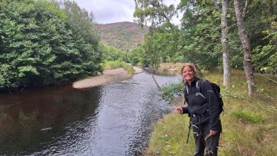 Georgia smiles wearing a rucksack in front of a river and hill