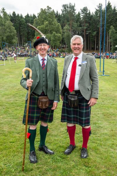 Two people in Highland dress standing in a field with a crowd if people far in the background.