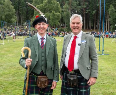 Two people in Highland dress standing in a field with a crowd if people far in the background.