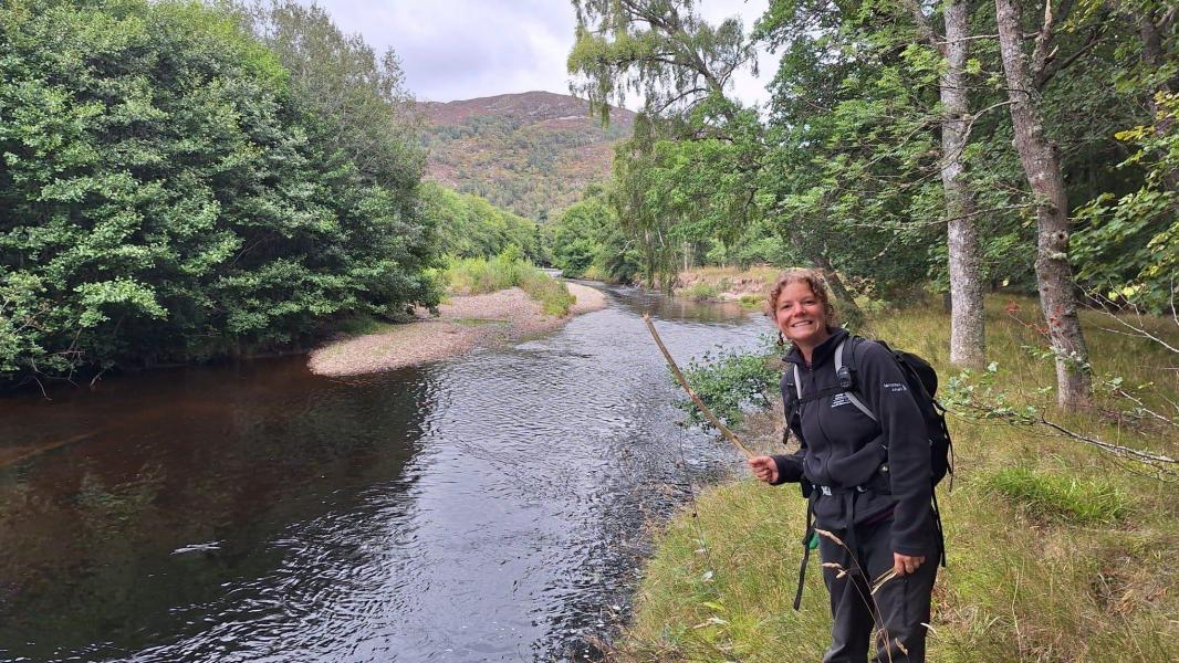 Georgia smiles wearing a rucksack in front of a river and hill