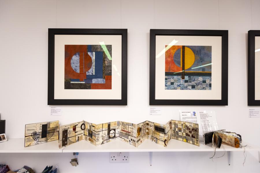 Two framed pieces of textile art hanging on a wall, with a shelf below holding folding paper collage artworks.