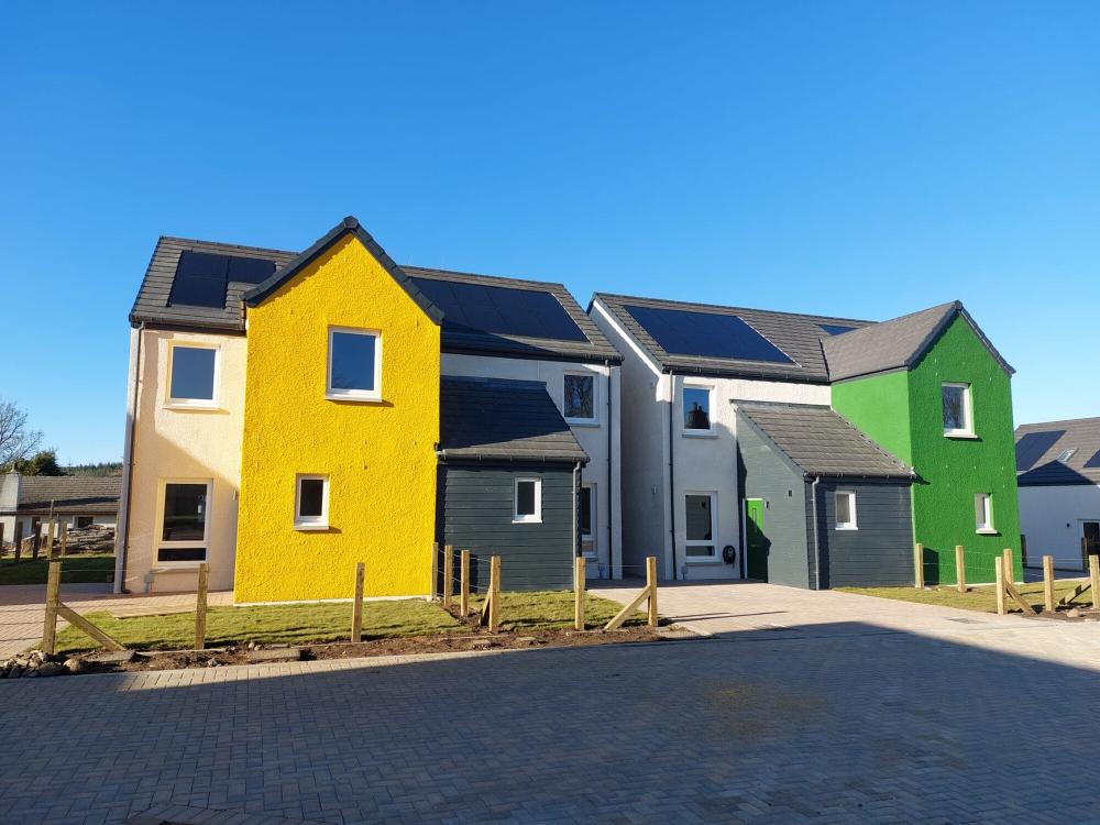 Affordable housing with bright coloured fronts and solar panels on roof, with blue sky behind them at Tomintoul