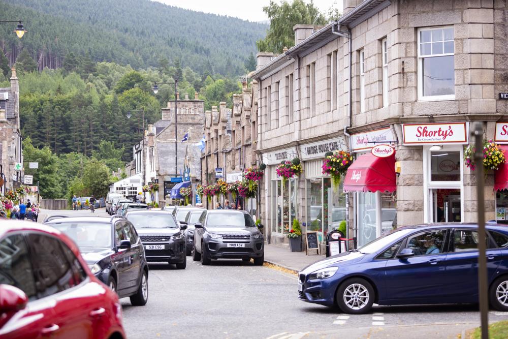 A view of a street in Ballater with cars, shops and wooded hills in the background
