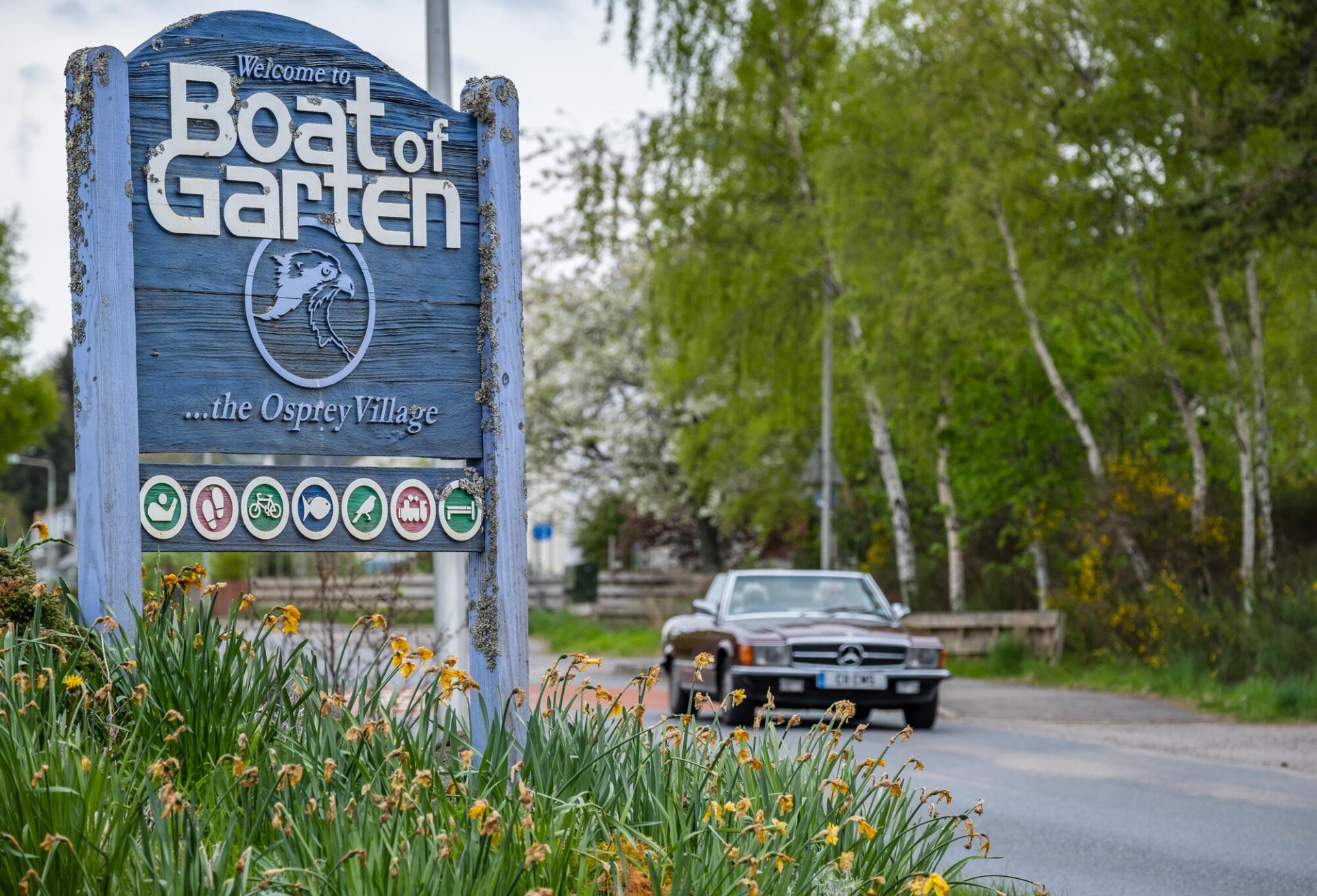 Welcome to Boat of Garten sign with car driving past and daffodils in the foreground.