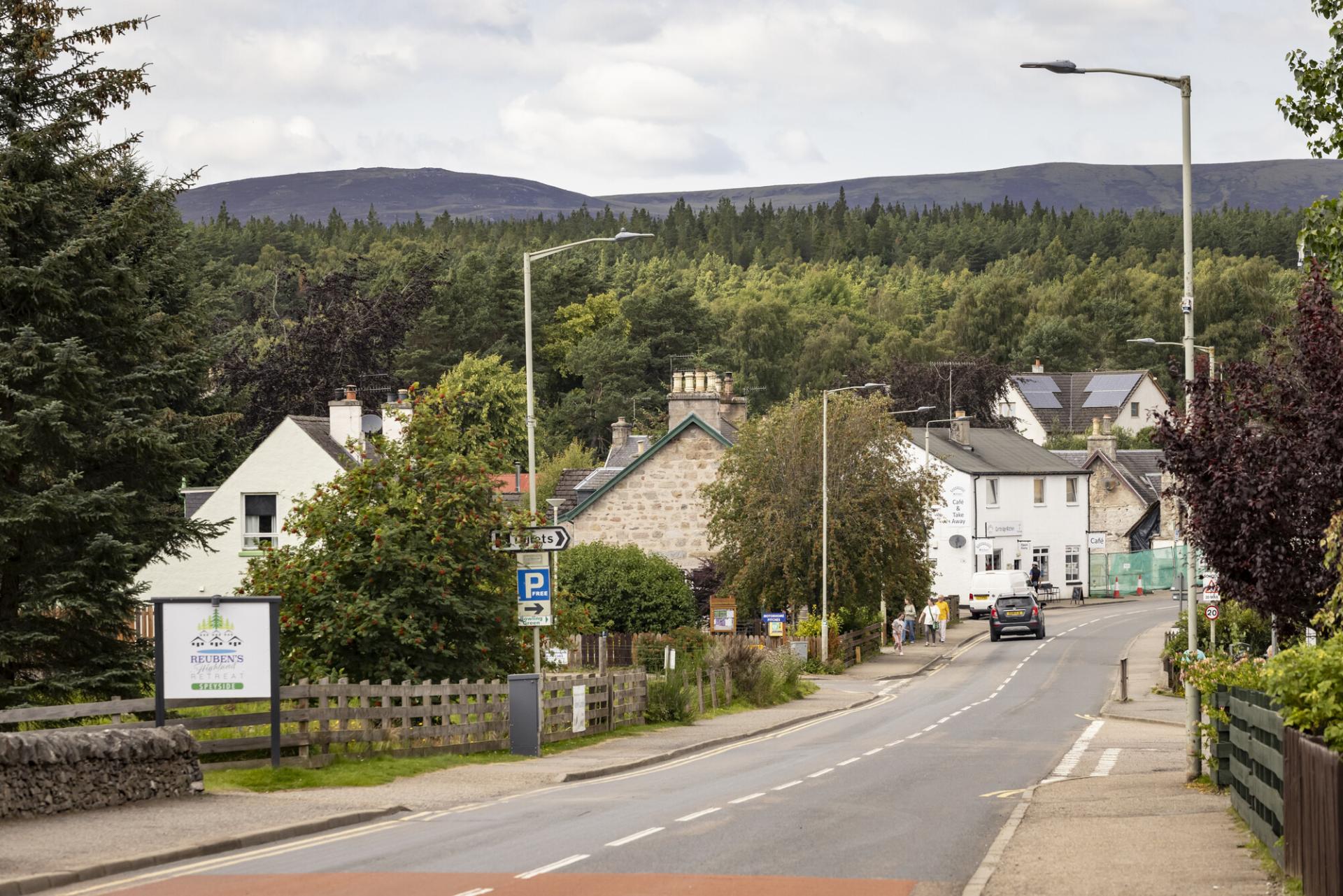 A view down a road in Carrbridge, with signs for holiday retreat, parking, toilets, and a cafe, with the woodland and hills in the background.