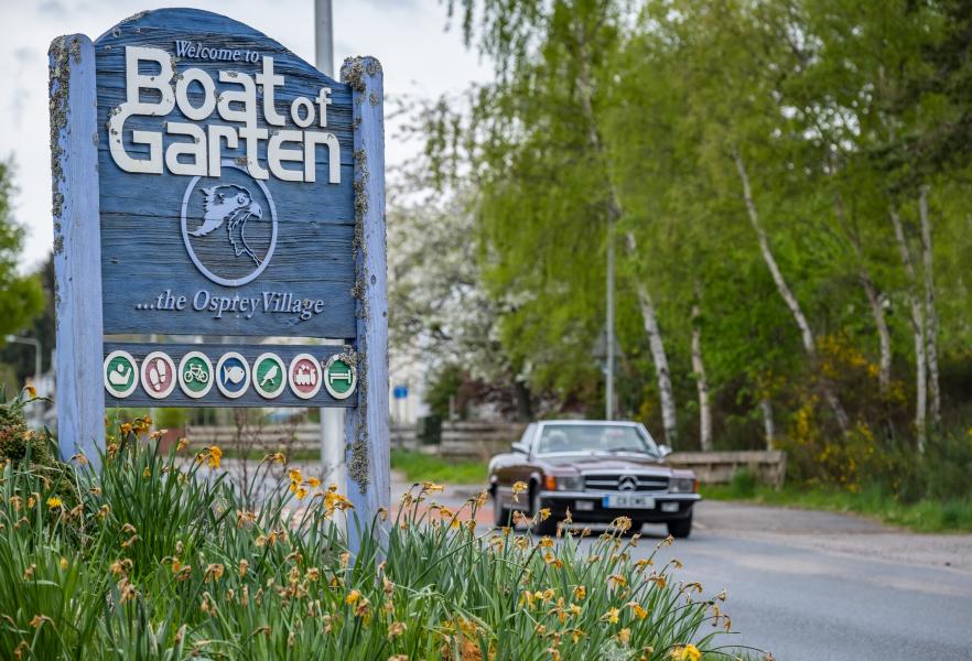 Welcome to Boat of Garten sign with car driving past and daffodils in the foreground.