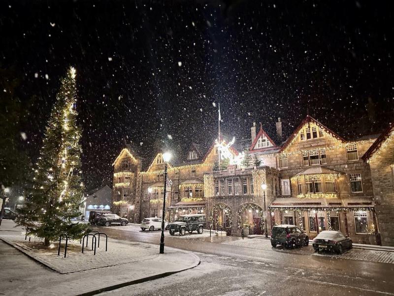 Snow falling at night in Braemar with a lit up hotel and tree.
