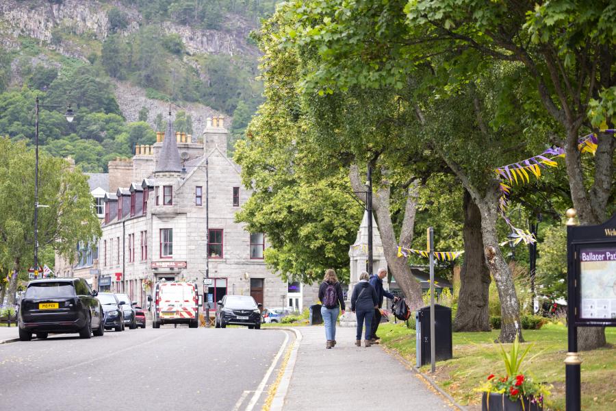 A street scene in Ballater adjacent to a village green with trees and bunting, a road with cars, shops in the background, and hillside