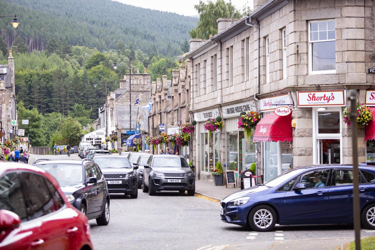 A view of a street in Ballater with cars, shops and wooded hills in the background