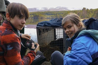 A young boy and girl beside a beaver in a crate at a loch with mountains in the background.