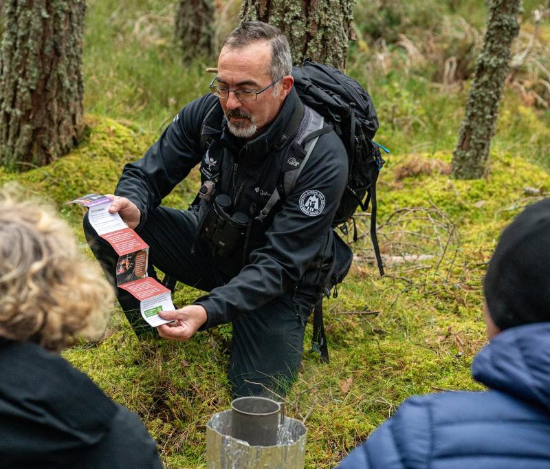 Ranger Duncan kneels with camping visitors on a mossy floor, with a camping stove in sight