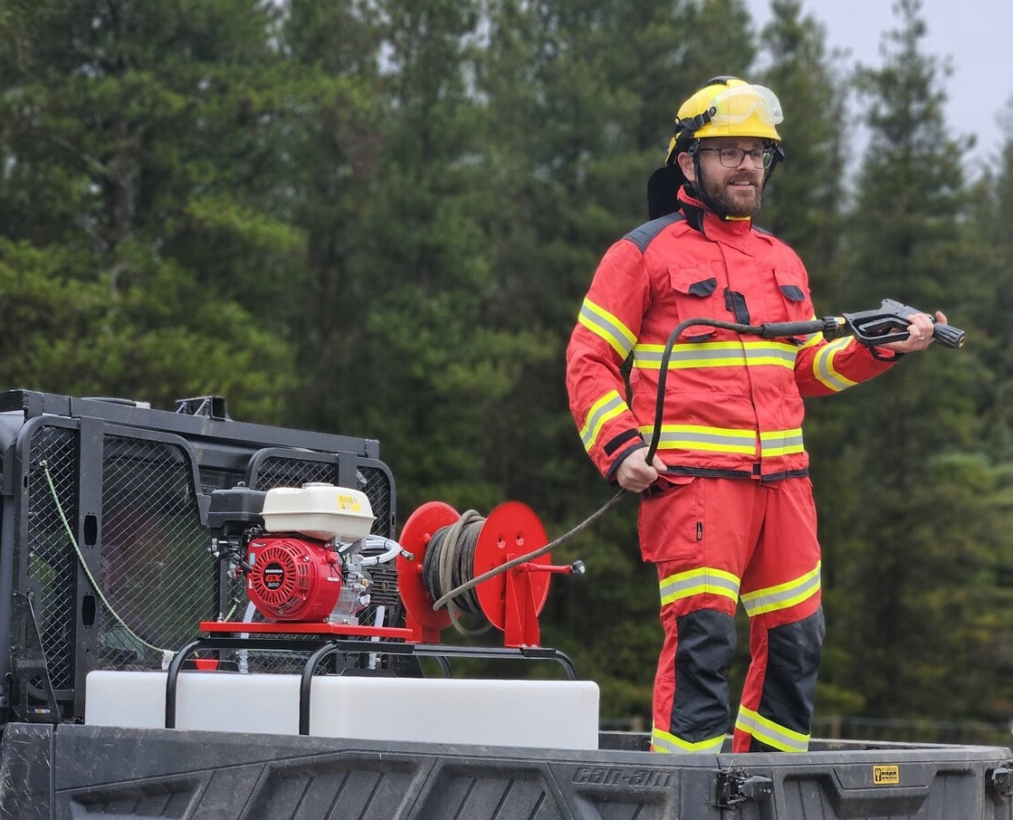 person standing on a vehicle wearing fire fighting PPE and holding a water hose