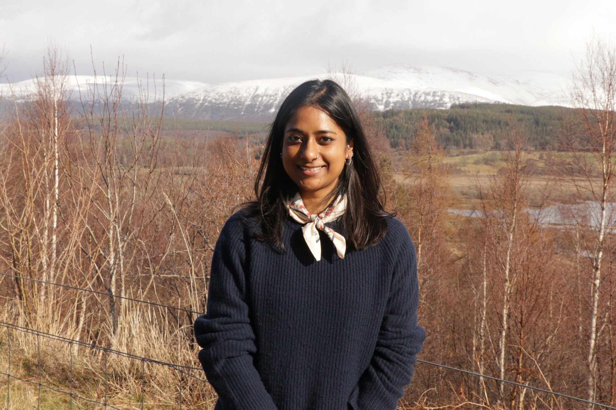 person in navy jumper and light coloured scarf standing in front of a landscape of autumn trees and snow-topped hills.