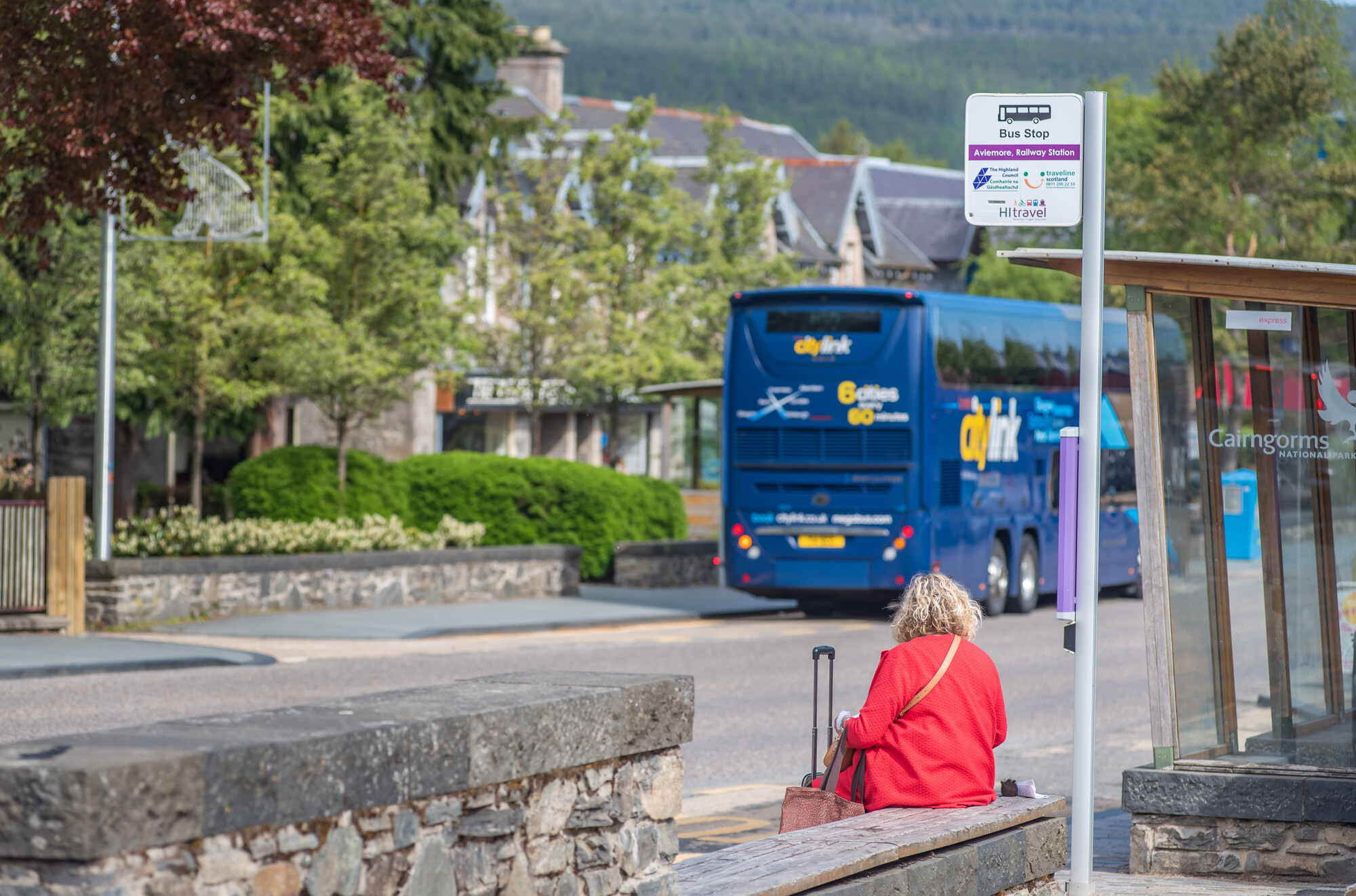 A woman waits for the bus with her luggage at a bus stop in Aviemore. There is a bus stopped at the bus stop across the street.