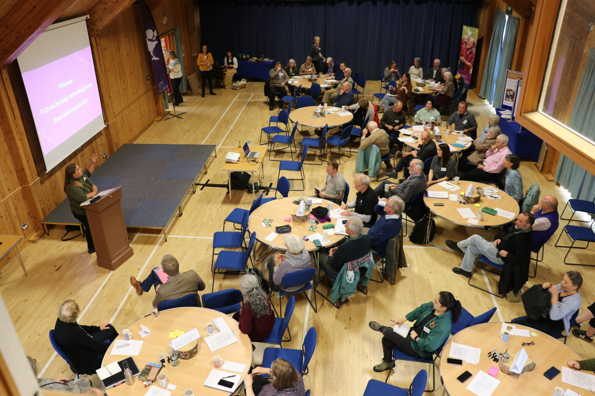 An audience sitting at tables in a hall listening to a speaker present in front of a screen.