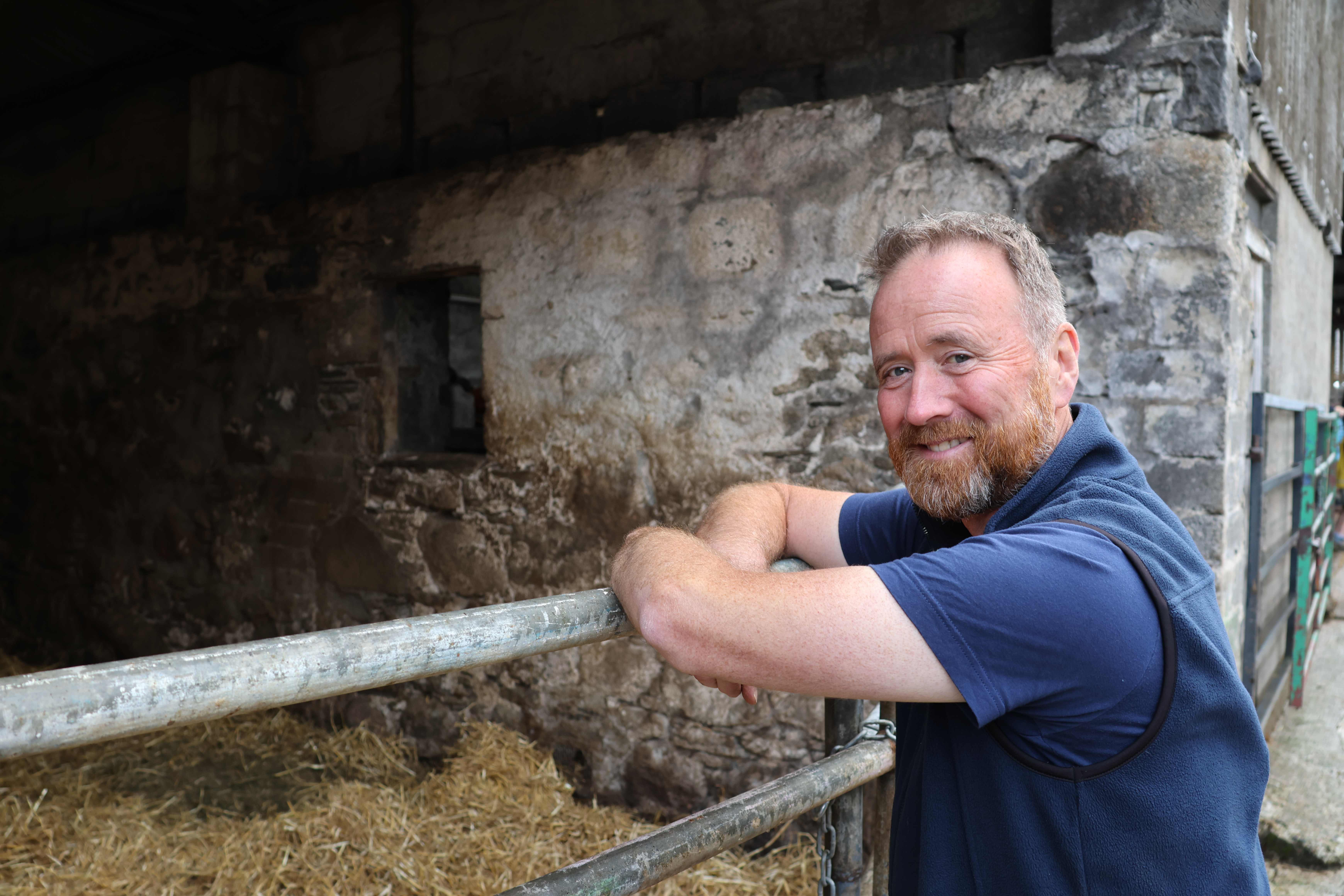 Man leaning on a gate to a cow shed, looking at the camera and smiling