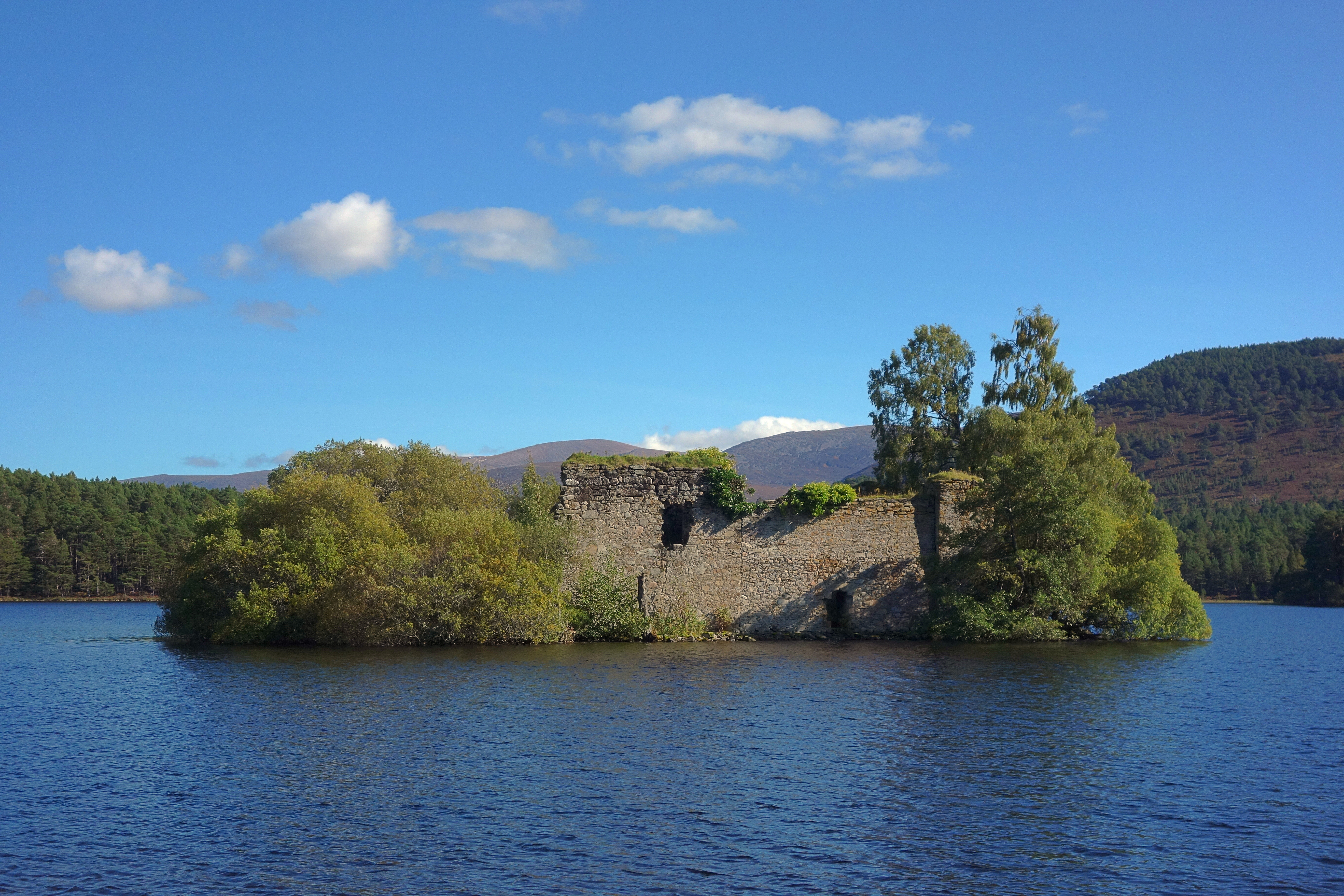 Image of a ruined castle surrounded by trees on an island in the middle of a loch, with woods and hills in the background.