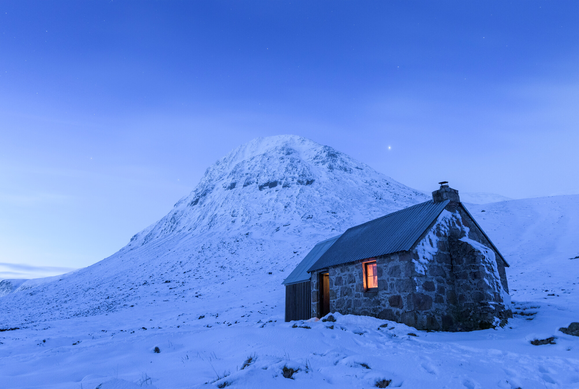 Bothy in a snow covered landscape with mountain in background.