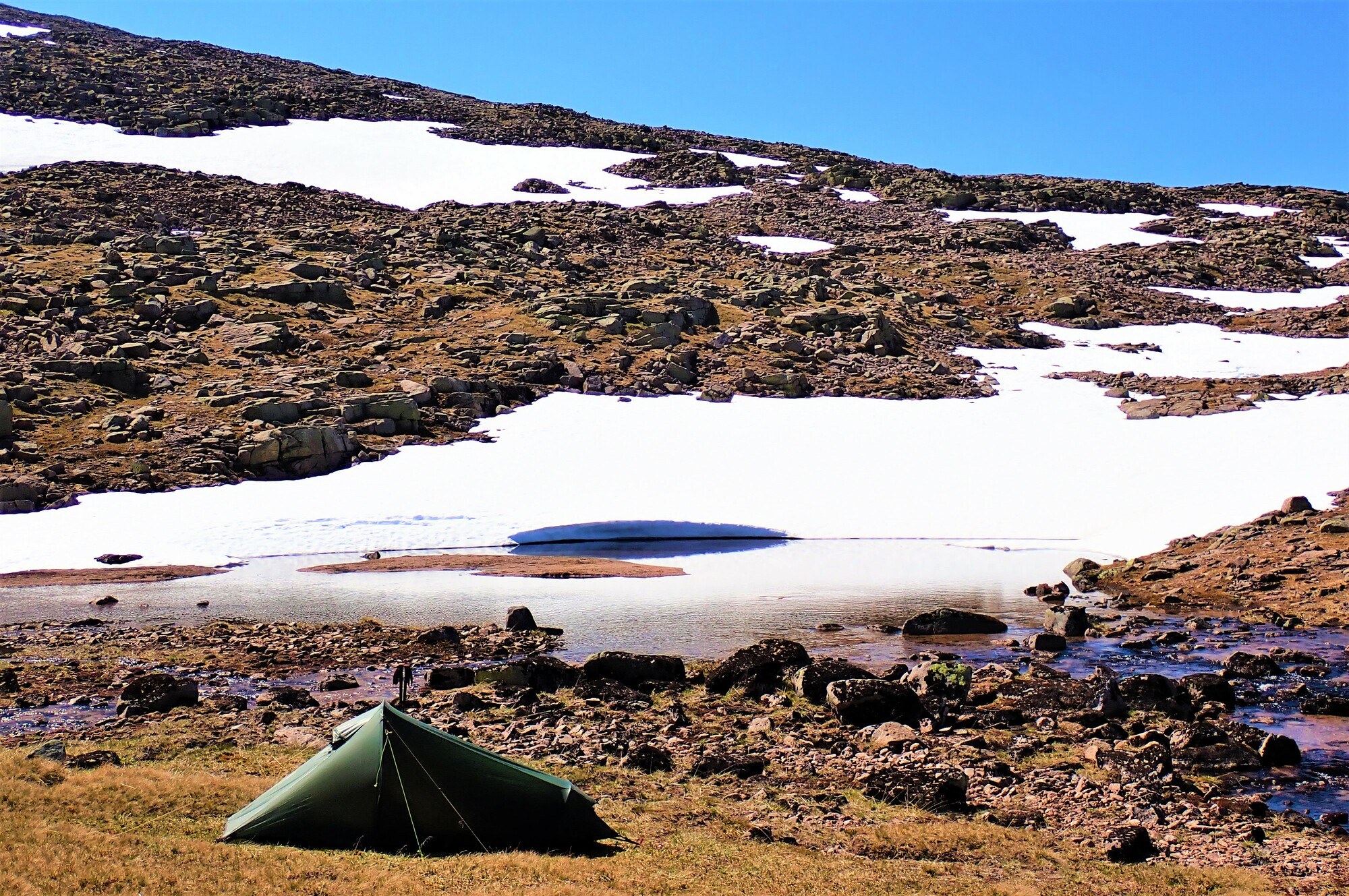 Wild camping in a small tent, next to a small lochan with snow in the background.