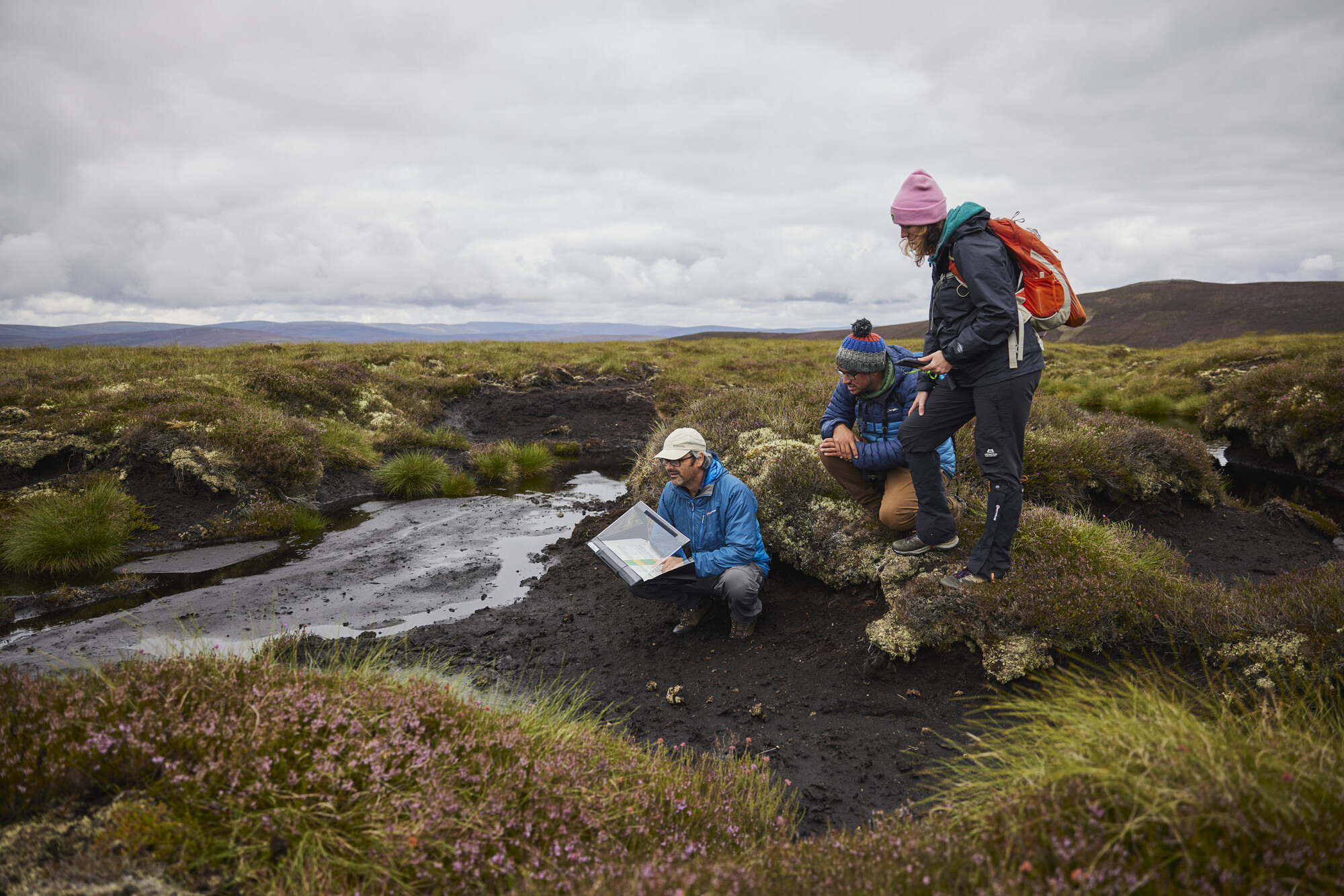 A woman in a mauve hat and waterproofs stands next to two men crouching next to a peat bog, one wearing a white cap and the other a multi-coloured bobble hat. One man makes notes in a waterproof folder.