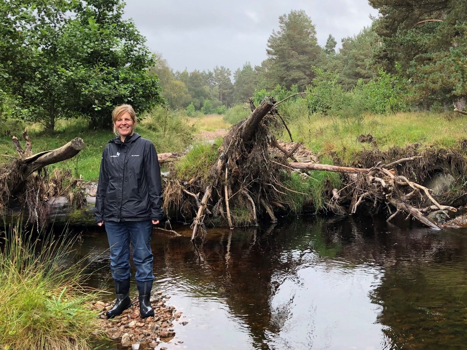 Image of Sally Mackenzie, wearing wellington boots and a black jacket, stood beside Allt Lorgy near Carrbridge.