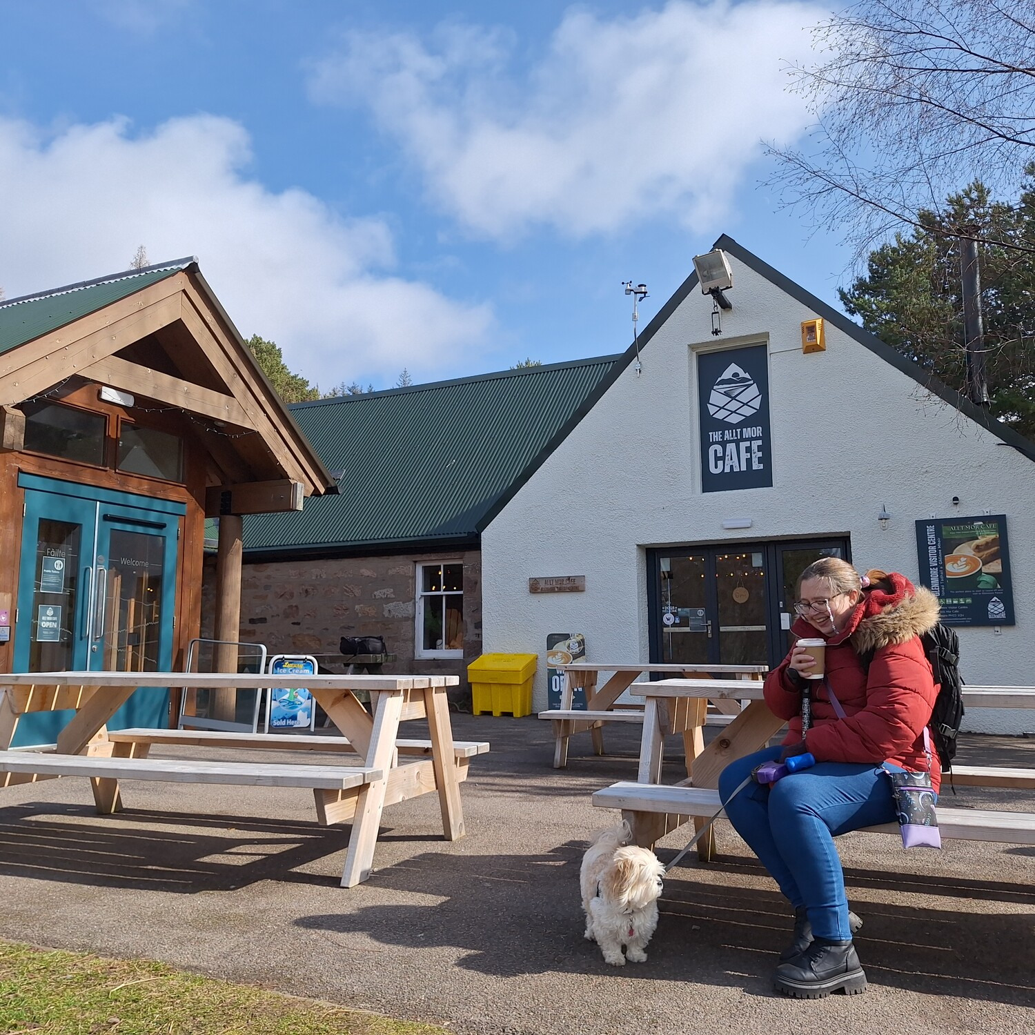 A visitor in a red jacket sits on a picnic bench outside Glenmore Visitor Centre with their white dog on a lead.