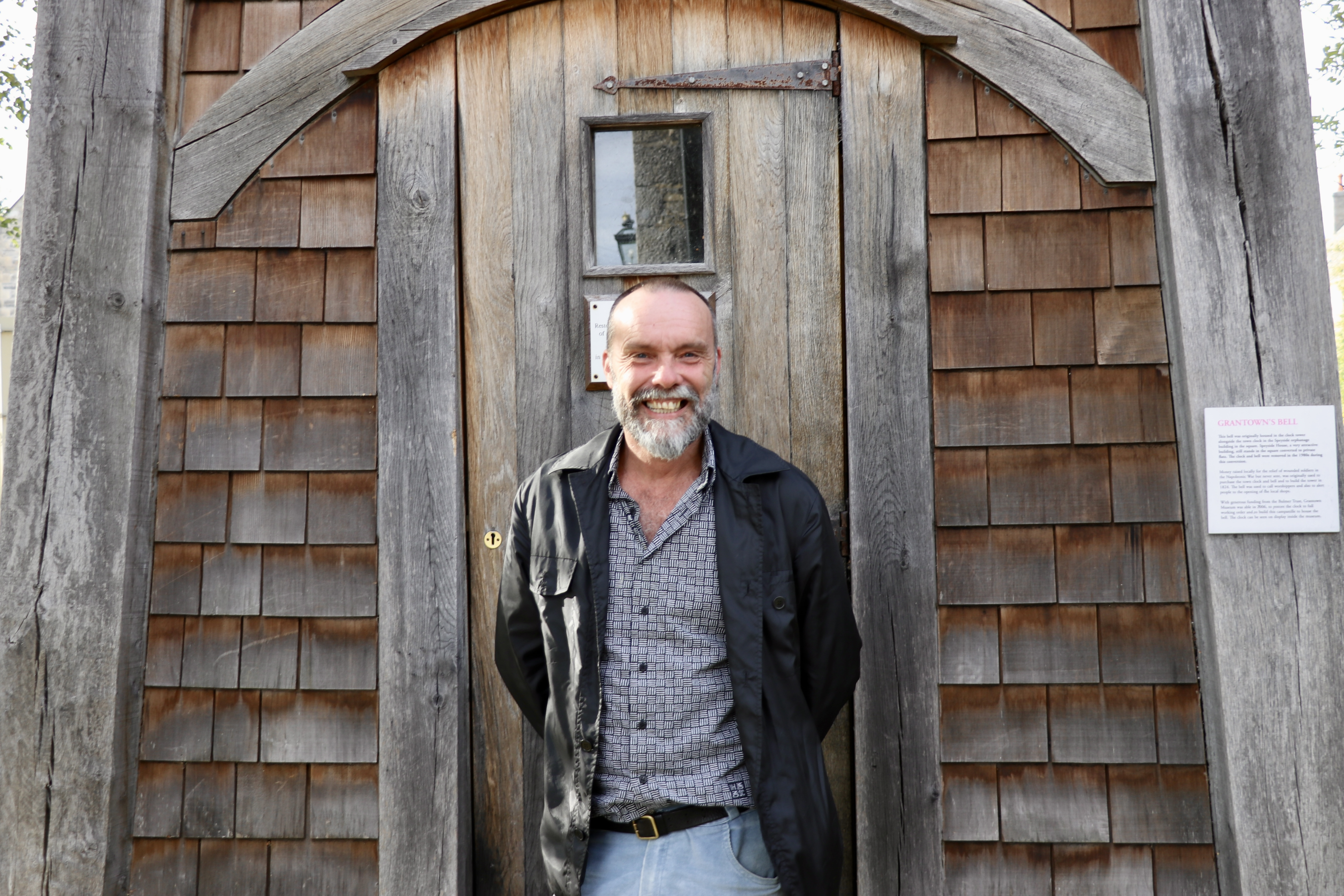 A smiling Dan Cottam stands in front of a wooden building with shingle tiles and a heavy wooden door.