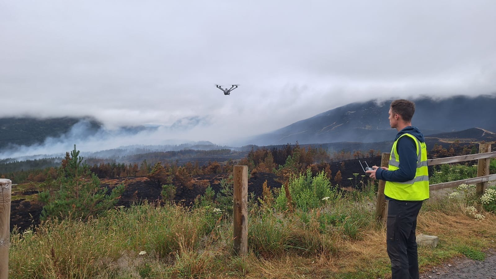 A thermal drone pilot in a yellow hi-vis vest looks out across the scene of the Glenmore wildfire