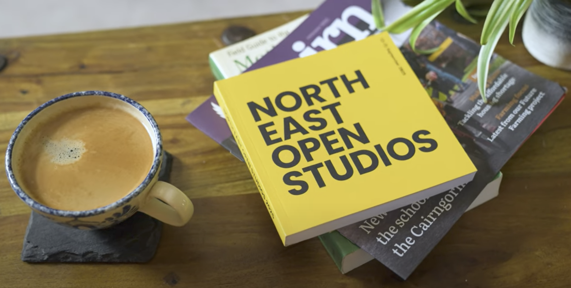 Image of a cup of coffee on a slate coaster with a pile of books and brochures to the right. At the top is the new North East Open Studios brochure in bright yellow.