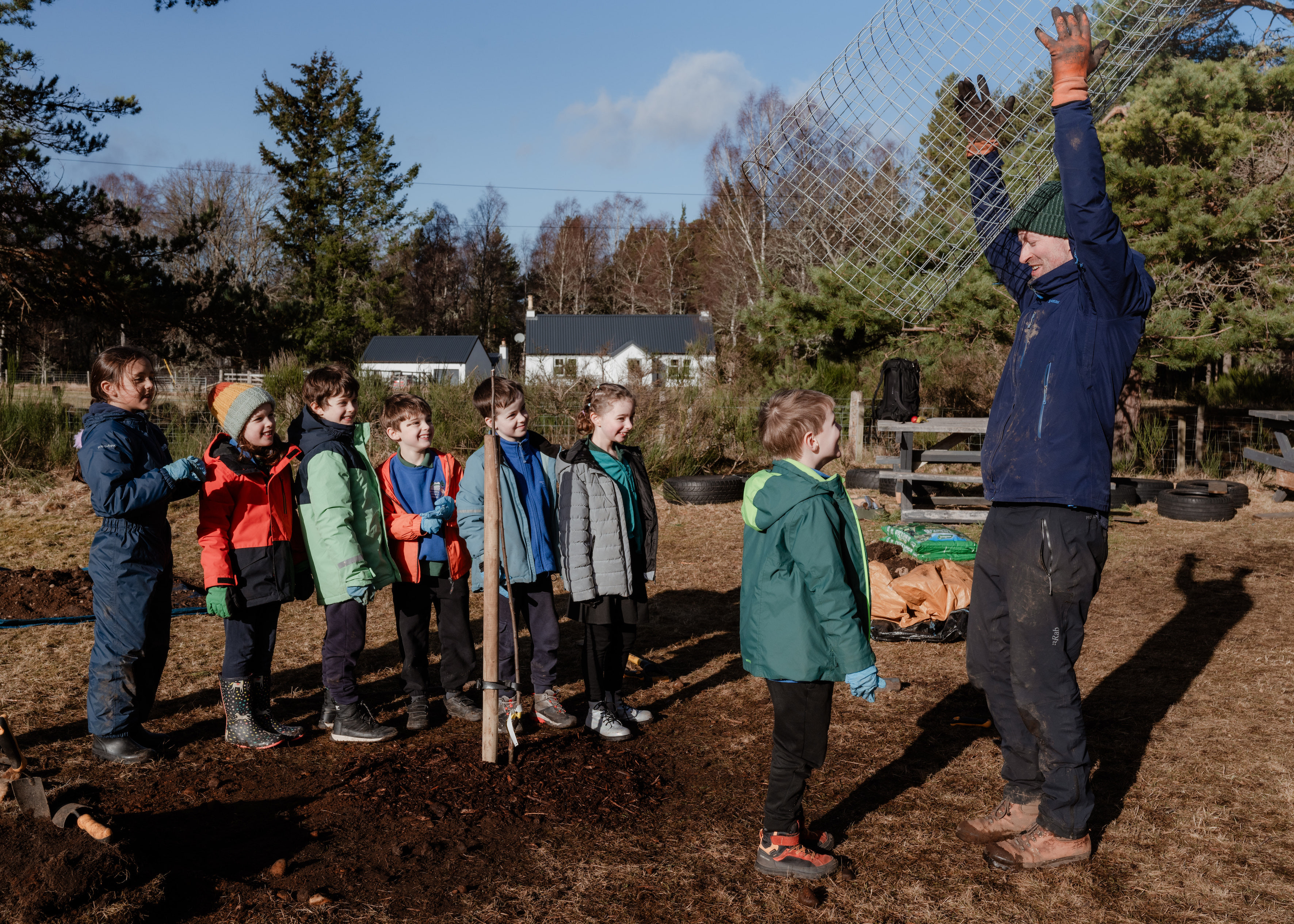 A group of children standing in a line with an adult on the right. All standing in a garden.