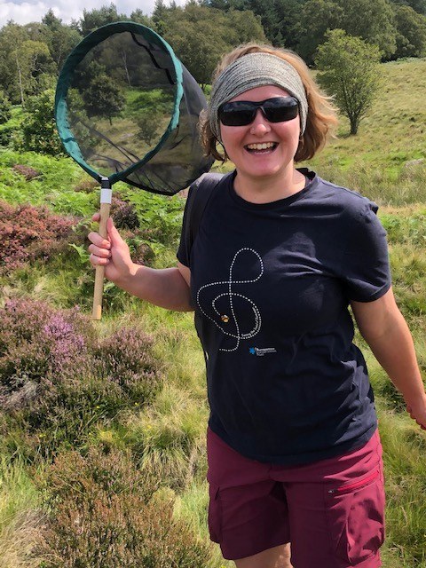 Image of Annie Ives, wearing shorts, t-shirt and sun glasses holding a short handled net in a grass and heater covered field.