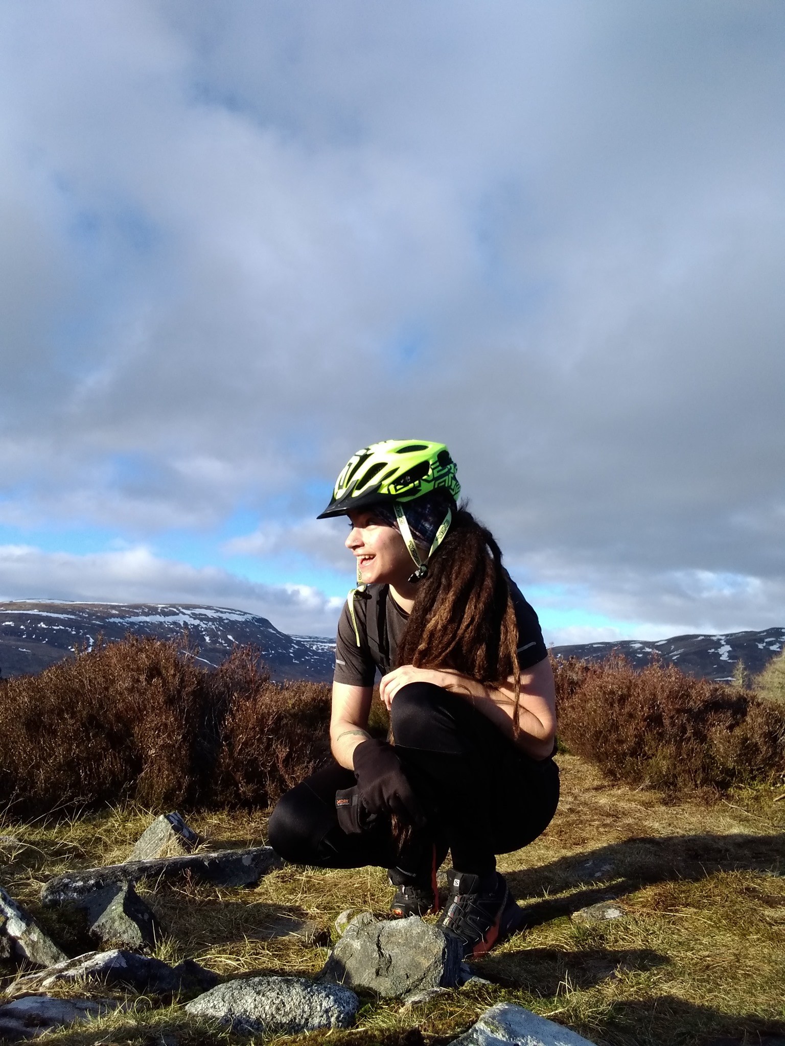 Image Bo Mansell (they / them), wearing a black t-shirt and trousers and a lime green cycle helmet, smiling whilst kneeling on stony / grassy ground surrounded by heather with a snowy mountain backdrop,