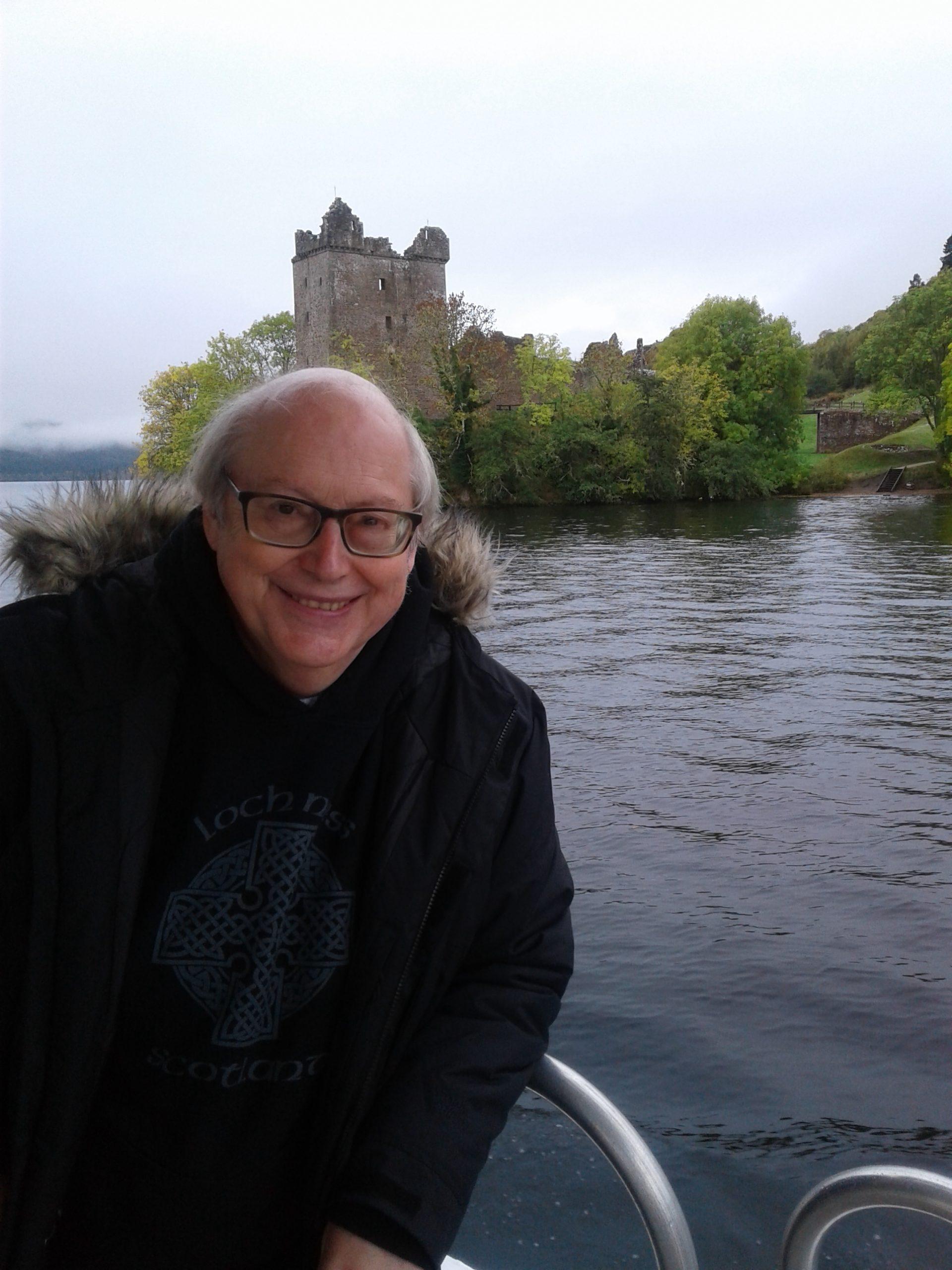 Image of Kevin Hutchens, wearing a jacket smiling at the camera whilst leaning on the side of a railing over water the ruins of a small castle on an island in the background.
