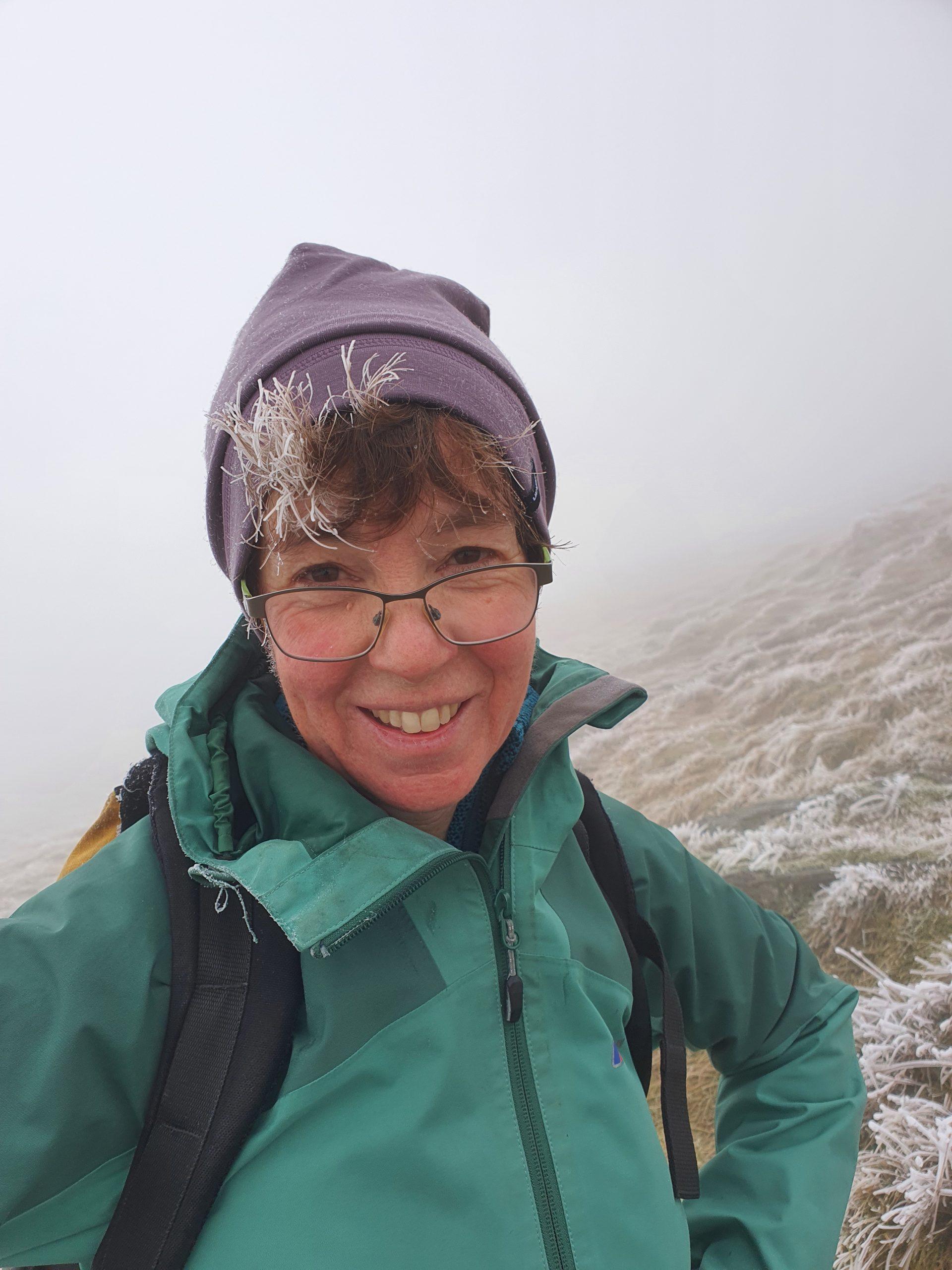 Image of Heather Earnshaw (she/her), wearing a green jacket and rucksack and a lilac woolly hat, smiling at the camera with a wintry hillside in the background.